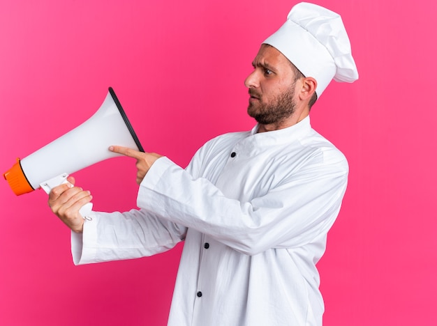 Fruncir el ceño joven cocinero de sexo masculino caucásico en uniforme de chef y gorra de pie en la vista de perfil sosteniendo y tocando el altavoz mirándolo aislado en la pared rosa