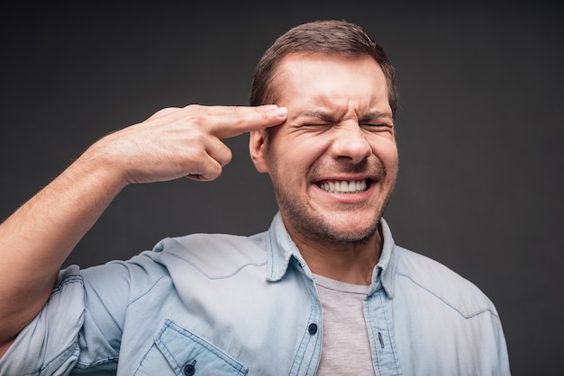 Frustração e desespero. Close de um jovem bonito gesticulando com uma arma e mantendo os olhos fechados em pé contra um fundo cinza