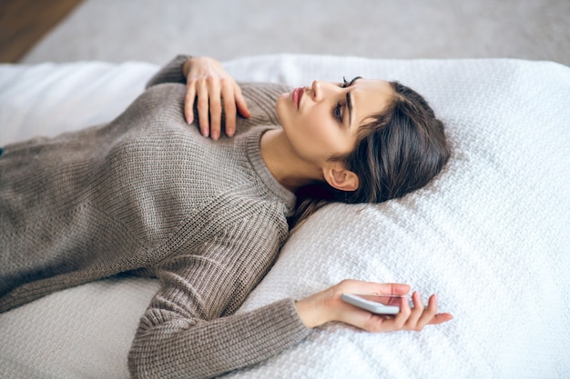 Frustración. Mujer joven de pelo oscuro acostado en la cama con un teléfono en la mano y mirando frustrado