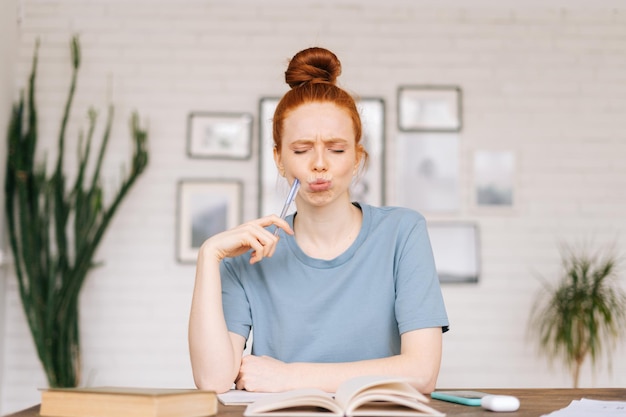 Frustrada y aburrida estudiante pelirroja sentada en una mesa con libros de texto y un libro de trabajo, mirando la cámara. Chica estudiante preparándose para los exámenes universitarios.