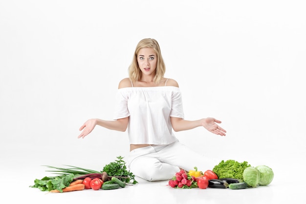 Frustrada mujer rubia extiende sus manos en ambas direcciones y muchas verduras frescas sobre un fondo blanco.