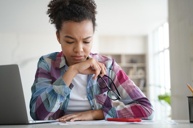 Frustrada niña afro está sobrecargada de trabajo estudiando a distancia en casa La colegiala está cansada de la tarea