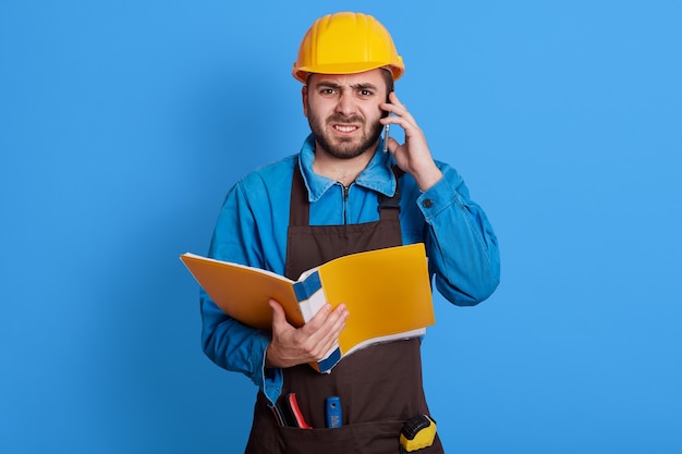 Frustrado capataz de construção falando com o funcionário em seu telefone celular, com expressão facial de raiva, vestindo uniforme azul, capacete amarelo e avental marrom, posando isolado na parede colorida.