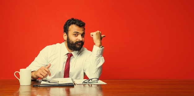 Frustrado joven empresario indio o asiático con barba presentando algo mientras está sentado en el escritorio de la oficina