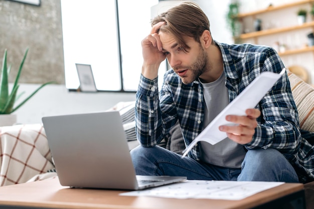 Foto frustrado y triste joven caucásico con ropa informal elegante, diseñador web independiente, director ejecutivo sentado en la sala de estar en el sofá trabajando en una computadora portátil en un proyecto, analiza documentos molestos con el resultado