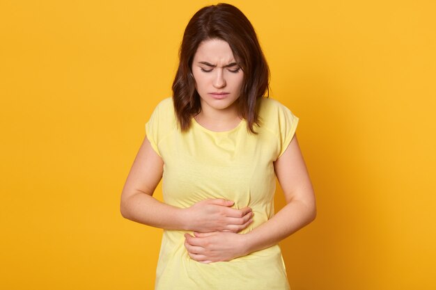 Frustrierte brünette Frau, die Hände auf Bauch hält, fühlt Schmerzen wie Menstruation oder Krankheit, trägt lässiges T-Shirt, bleibt zu Hause