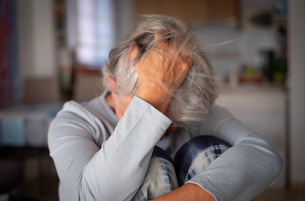 Frustrierte depressive alte Frau, die das Gesicht mit der Hand in den Haaren bedeckt und allein zu Hause auf der Couch sitzt