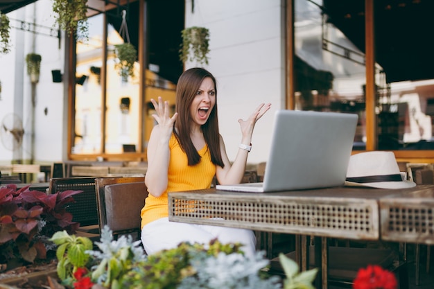 Frustrierte Frau im Straßencafé im Freien, die am Tisch mit einem modernen Laptop-PC sitzt und in der Freizeit die Hände im Restaurant ausbreitet