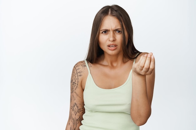 Foto frustrierte junge frau, die hand schüttelt und verwirrt in die kamera schaut, tanktop trägt und vor weißem hintergrund steht