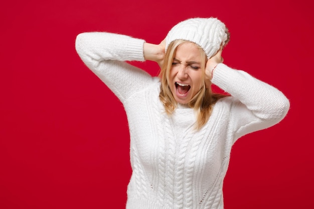 Foto frustrierte junge frau im weißen pullover, hut isoliert auf rotem hintergrund im studio. gesunder modelebensstil, konzept der kalten jahreszeit. mock-up-kopienbereich. schreien, augen geschlossen halten, hände auf den kopf legen.