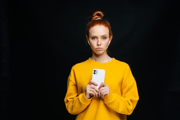 Frustrierte junge Frau mit Handy und Blick in die Kamera auf isoliertem schwarzem Hintergrund