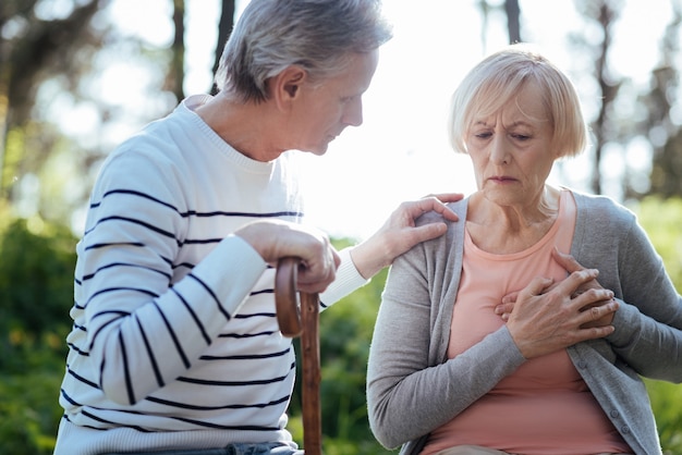 Frustrierte schwache alte Frau, die ihre Brust berührt und einen Herzinfarkt hat, während ihr alter Ehemann sich Sorgen um sie macht und draußen auf der Bank sitzt
