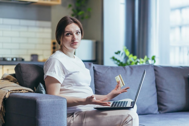 Frustrierte schwangere Frau versucht, zu Hause online einzukaufen, schaut frustriert auf die Kamera und verwendet einen Laptop mit einer Bankkreditkarte