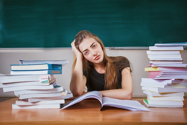 Frustrierte Studentin, die am Schreibtisch mit einem enormen Stapel von Studienbüchern im Klassenzimmer sitzt