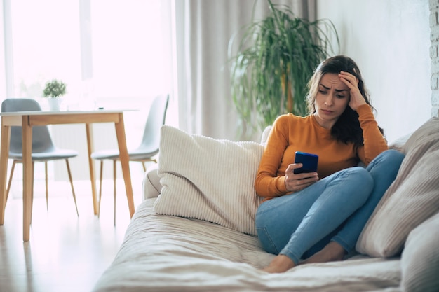 Frustrierte und deprimierte junge brünette Frau weint mit einem Smartphone in der Hand, während sie zu Hause auf der Couch sitzt