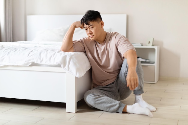 Frustrierter chinesischer Mann, der im Schlafzimmer auf dem Boden sitzt