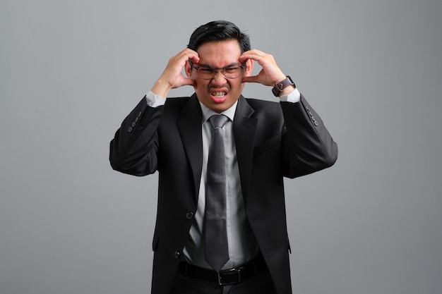 Frustrierter gutaussehender junger Geschäftsmann im formellen Anzug, der unter Kopfschmerzen leidet, weil Migräne auf grauem Hintergrund isoliert ist