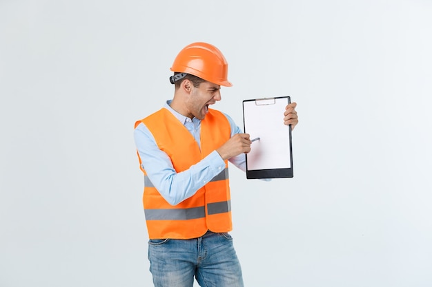 Frustrierter junger Ingenieur mit Bauarbeiterhelm und reflektierender Weste, der Fehler im Dokument auf grauem Hintergrund überprüft.