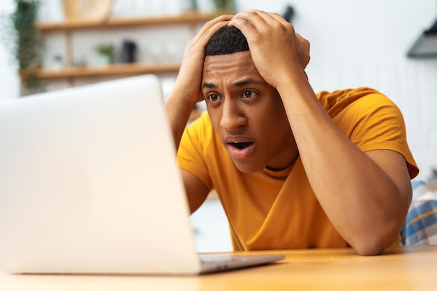 Frustrierter, trauriger afroamerikanischer Mann berührt den Kopf mit einem Laptop, hat Geld verloren, hat einen Fehler gemacht.