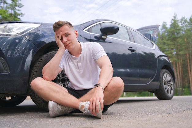 Frustrierter, trauriger, verärgerter Fahrer sitzt nach einem Verkehrsunfall in der Nähe eines kaputten Autos