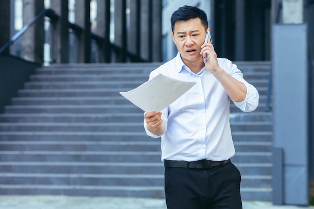 Frustrierter und wütender asiatischer Geschäftsmann, der am Telefon spricht und einen Brief und Dokumente von einer Bank außerhalb des Büros hält