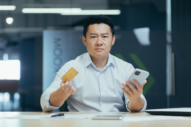 Frustrierter und wütender asiatischer Mann im Büro Geschäftsmann blickt in die Kamera und hält Telefon und Bank in der Hand