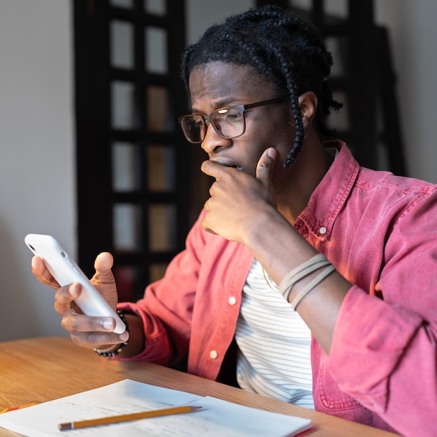 Frustrierter, verängstigter Afroamerikaner sitzt mit Handy am Tisch, das den Mund mit der Hand bedeckt