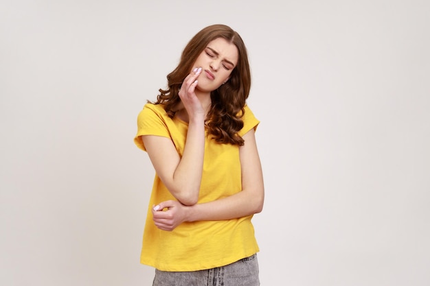 Frustriertes, gewelltes Teenager-Mädchen mit gelbem, lässigem T-Shirt, das die Wange berührt und vor Schmerzen zusammenzuckt und sich schreckliche Zahnschmerzen bei Parodontitis verspürt Studioaufnahme isoliert auf grauem Hintergrund