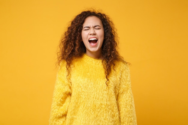 Frustriertes junges afrikanisch-amerikanisches Mädchen im Pelzpullover posiert isoliert auf gelb-orangefarbenem Wandhintergrund, Studioportrait. People-Lifestyle-Konzept. Mock-up-Kopienbereich. Augen geschlossen halten, schreien.