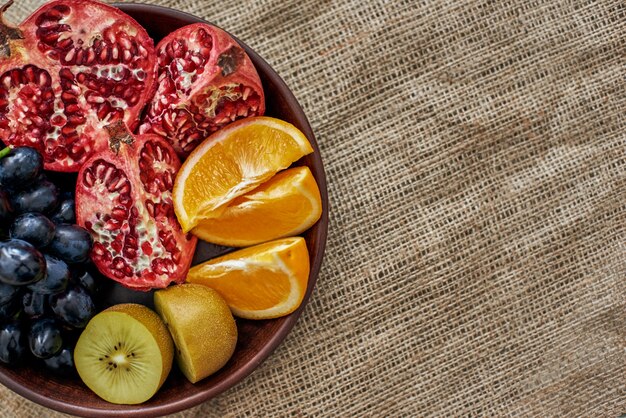 Fruta jugosa. Fotografía cenital de un plato lleno de jugosas frutas en rodajas en un plato de madera con uvas, naranja, granada y kiwi en la tela de arpillera