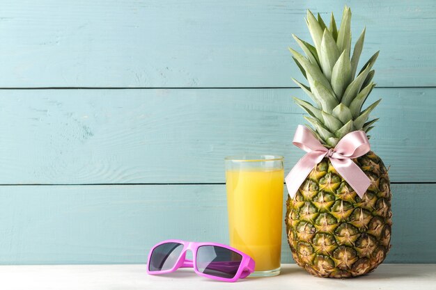 Fruta madura do abacaxi com um laço rosa, óculos escuros e suco de abacaxi em uma mesa de madeira azul. lugar para texto.