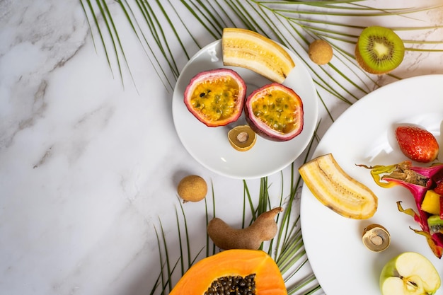 Fruta de la pasión en rodajas, longan, plátano en un plato blanco. Frutas exóticas y hojas tropicales sobre fondo de mármol blanco con espacio para copiar el texto. Comida, concepto de verano. Vista superior.