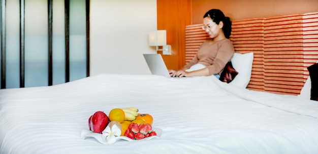 Frutas en la bandeja frente a los turistas utilizan la computadora portátil en la cama en la habitación del hotel de lujo.