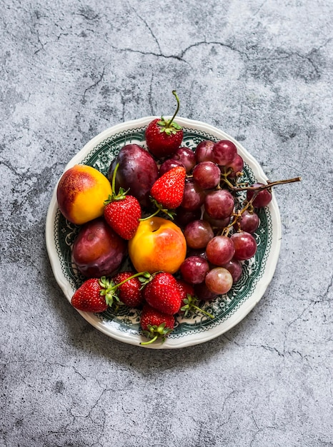 Frutas y bayas de verano melocotones ciruelas fresas uvas en un plato vintage en una vista superior de fondo gris