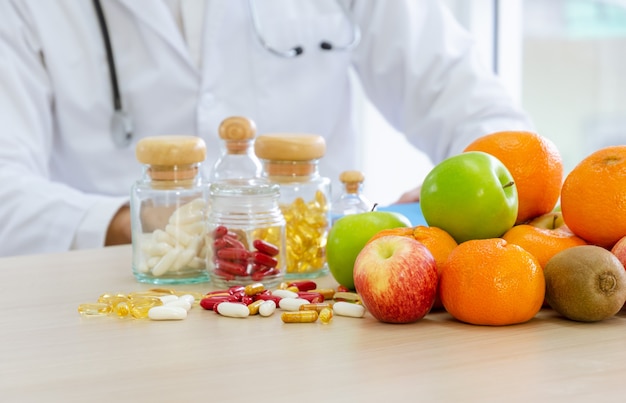 Frutas frescas, cápsulas e vitaminas na mesa. especialista nutricional com estetoscópio em volta do pescoço desenvolvendo plano alimentar para melhorar os hábitos alimentares do paciente