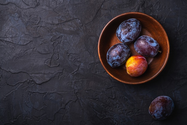 Frutas frescas de ciruela dulce en tazón de madera marrón, fondo de textura negra, espacio de copia de vista superior