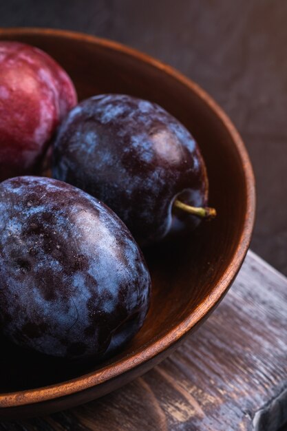 Frutas frescas de ciruela dulce en un tazón de madera marrón sobre la vieja tabla de cortar, mesa con textura negra, vista de ángulo macro