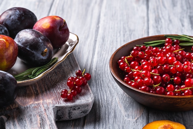 Frutas frescas de ameixa doce inteiras e fatiadas em um prato com folhas de alecrim na velha tábua de corte com bagas de groselha em uma tigela de madeira, superfície de madeira cinza, vista angular de perto