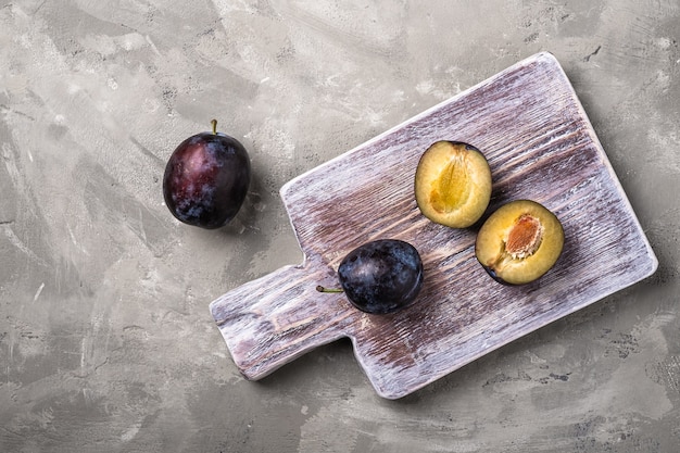 Frutas frescas de ameixa madura inteiras e fatiadas em uma tábua de madeira, fundo de pedra e concreto, vista superior