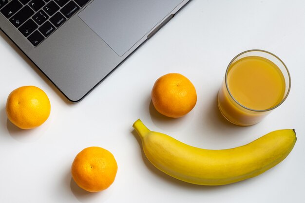 Frutas no local de trabalho. portátil, banana, mandarino e suco de laranja na mesa branca.