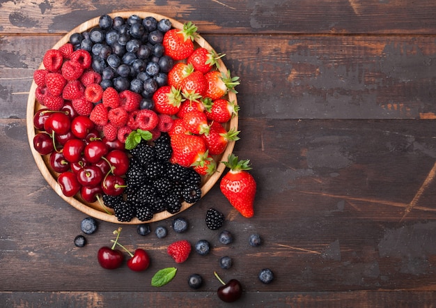 Frutas orgânicas frescas no verão se misturam em uma bandeja de madeira redonda no fundo da mesa de madeira escura. framboesas, morangos, mirtilos, amoras e cerejas. vista do topo