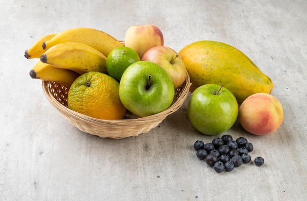 Frutas surtidas en una canasta sobre mesa gris claro.