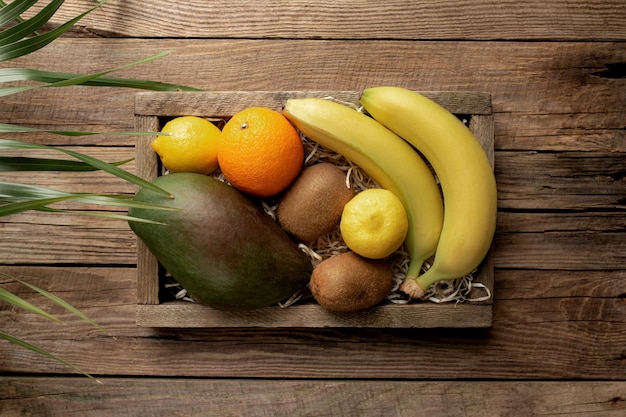 Frutas tropicales frescas en una caja de entrega de madera sobre una mesa de madera. Naranja, plátano, mango, kiwi y limón, vista superior.