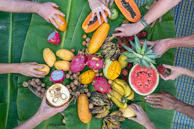 Foto frutas tropicales en hojas de plátano verde y manos de personas. grupo de amigos felices con buena comida, disfrutando de la fiesta y la comunicación. mango, papaya, pitahaya, plátano, sandía, piña y manos