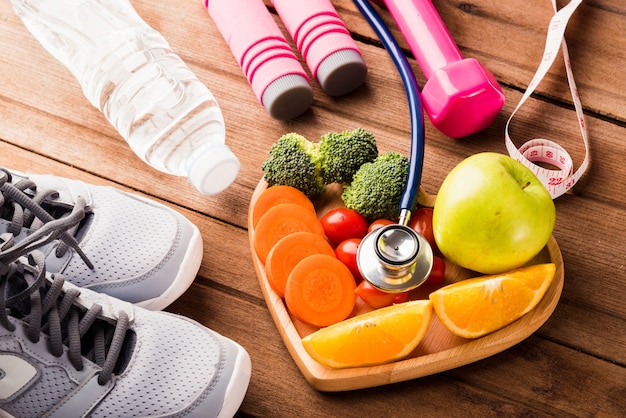 frutas y verduras frescas en madera de placa de corazón y equipo deportivo y estetoscopio médico