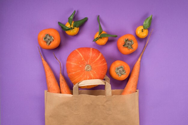 Frutas y verduras naranjas en bolsa de papel artesanal sobre fondo violeta.