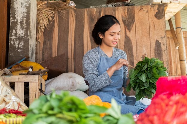 Frutería asiática mujer sentada sosteniendo espinacas para atar en el puesto de verduras