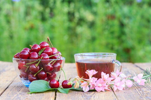 Frutos rojos de cereza madura con pedúnculos en un tazón de vidrio, taza de té y flores.