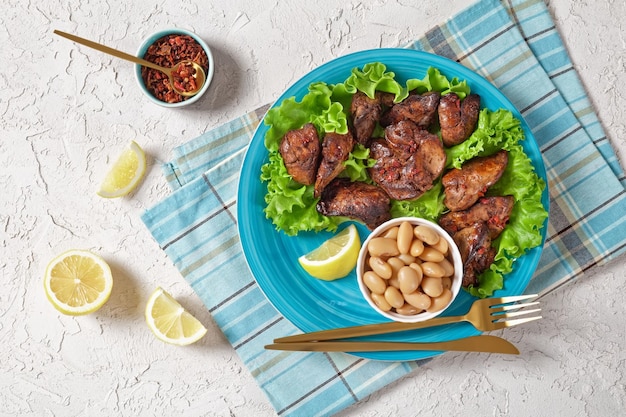 Fry de hígado de pollo Kaleji Fry servido en hojas de lechuga con frijoles de mantequilla y gajos de limón en un plato azul sobre una mesa blanca con textura cocina india plana