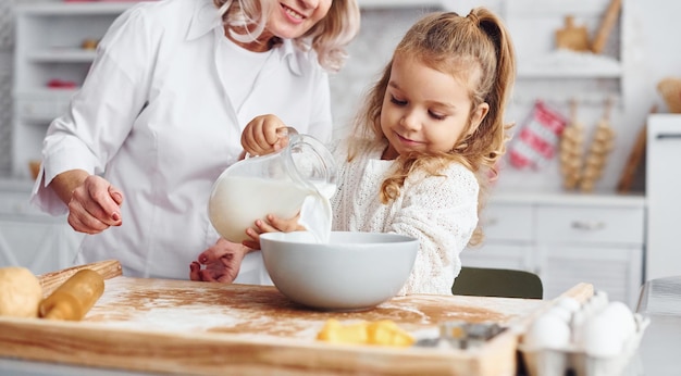 Füllt sich mit Milch Ältere Großmutter mit ihrer kleinen Enkelin kocht Süßigkeiten für Weihnachten in der Küche